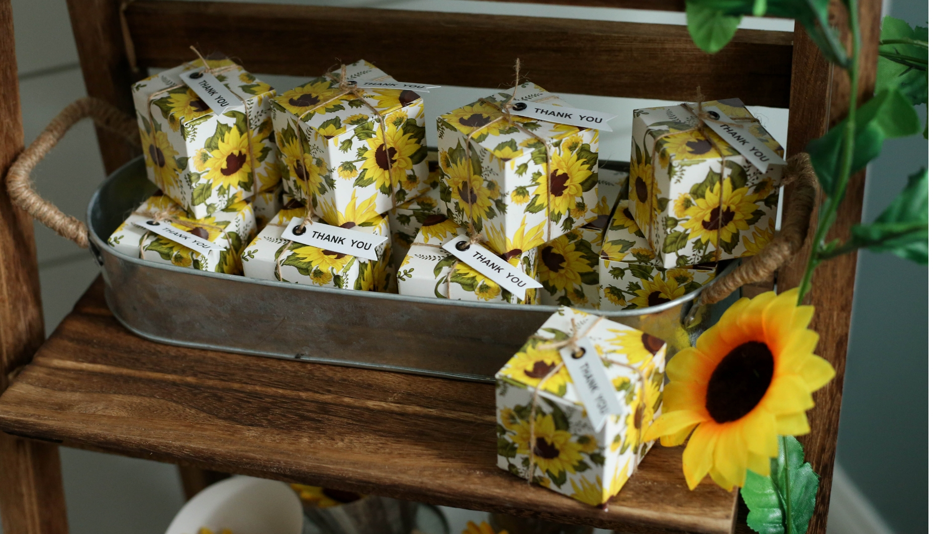Sunflower themed favor boxes are tied with jute twine and thank you tags, arranged in a metal galvanized tray with handles for a sunflower themed wedding dessert display on a ladder shelf