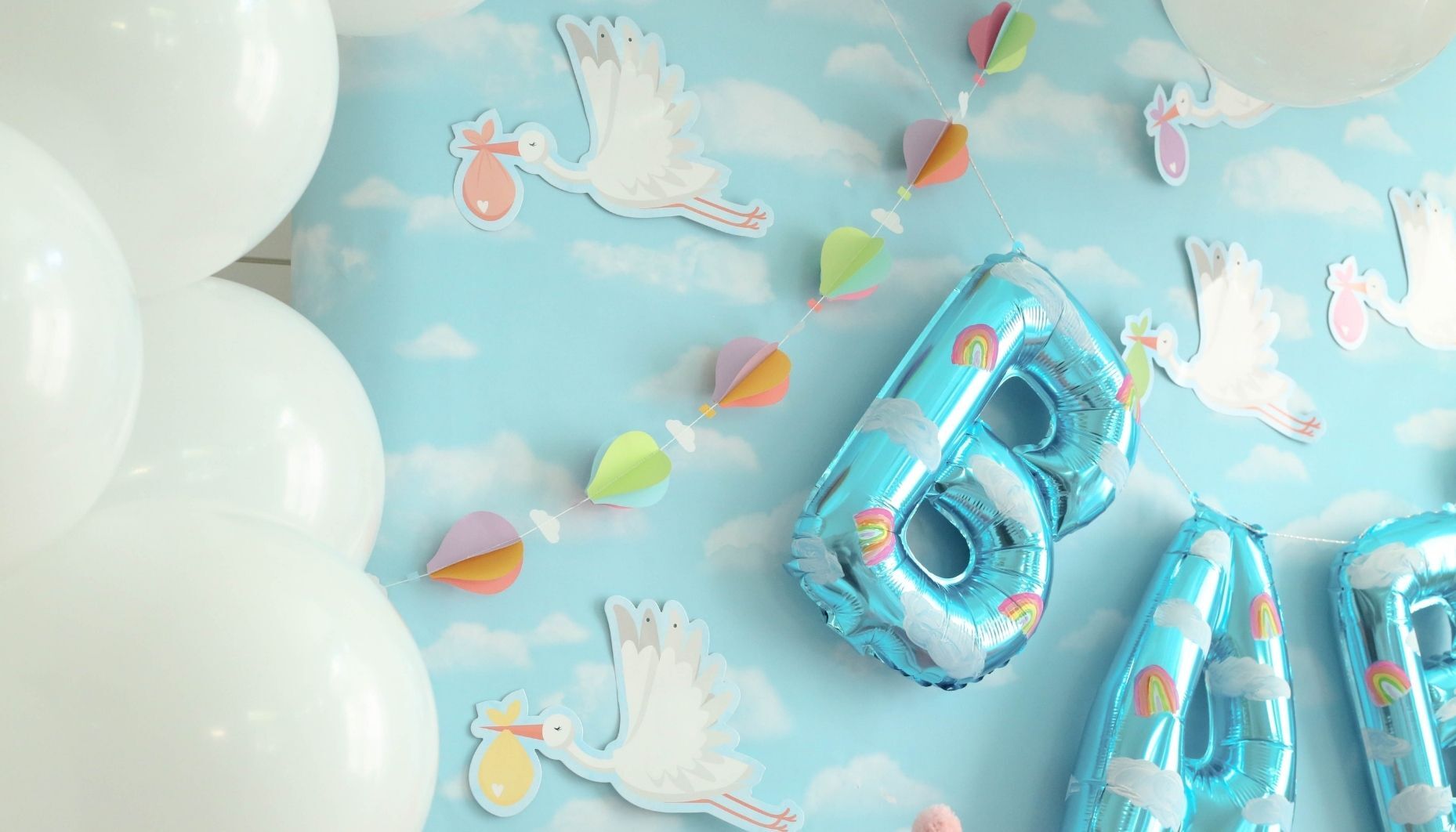 Hand painted mylar balloons have bright rainbows. A 3D hot air balloon garland and paper storks are hung with a white balloon garland on a cloud backdrop.