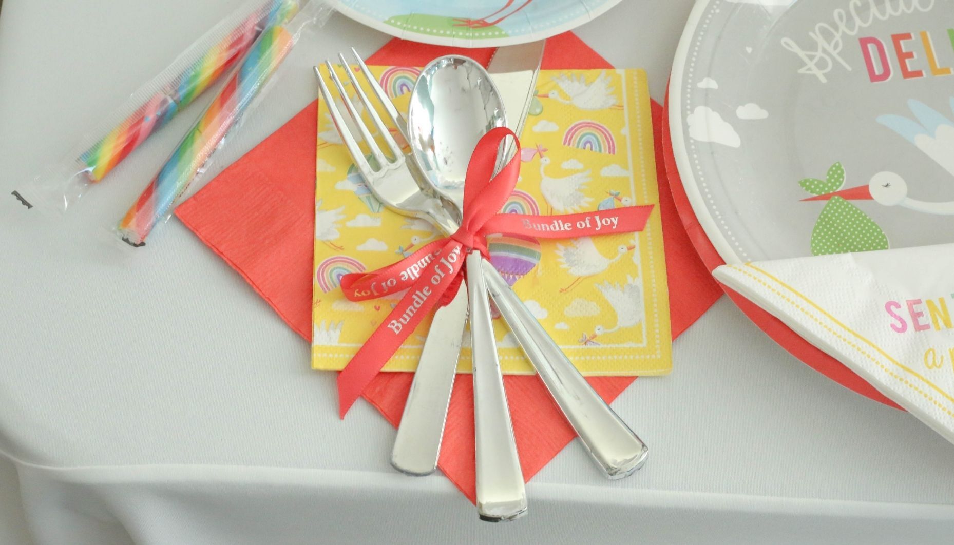 Customized coral satin ribbon reads, Bundle of Joy, and is used to tie silver flatware bundles together for a stork themed baby shower.